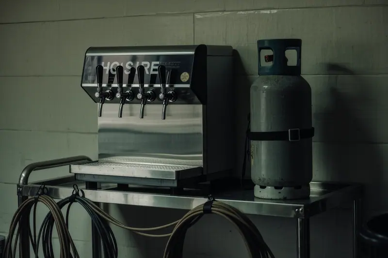 A hire beer dispense unit with two taps, a gas bottle and coiled dispense lines on a stainless steel trolley in a store room.