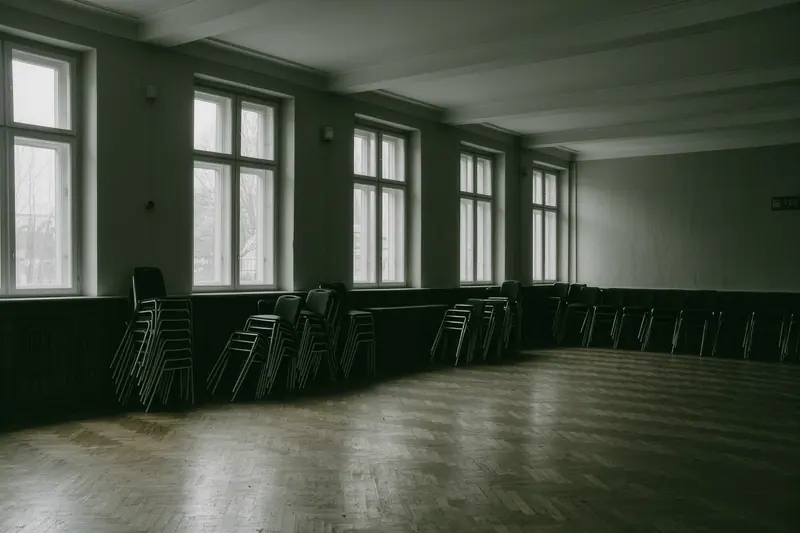 An empty hired hall with a parquet floor, stacked chairs against one wall and tall windows along the far side.