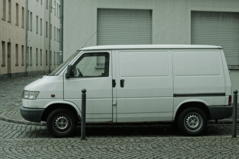 A small white van parked on a narrow cobbled side street lined with bollards and closed shutters.
