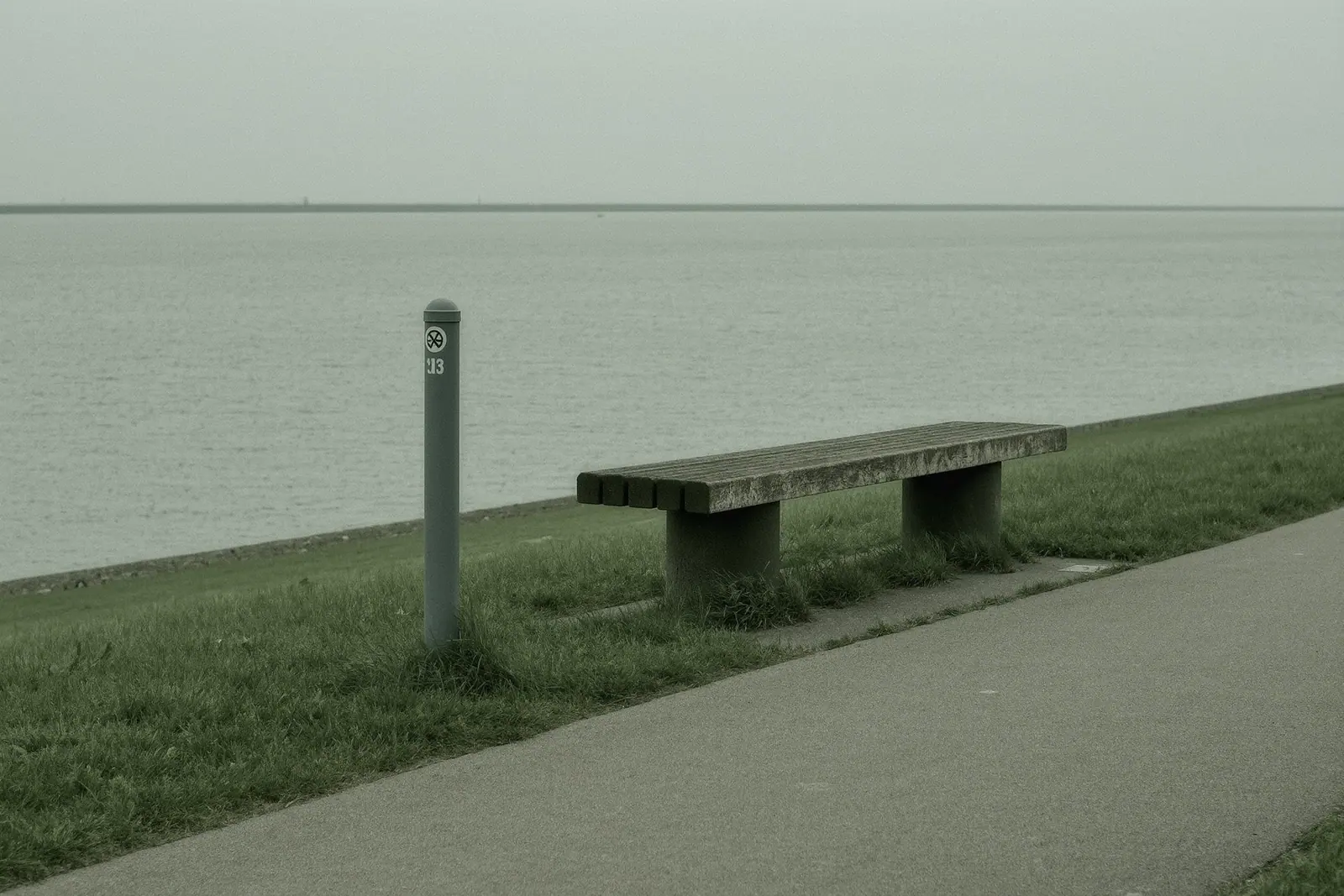 A paved cycle path along a river embankment with a plain metal route marker post and a weathered wooden bench in the foreground, grass bank and flat grey water behind, no riders.